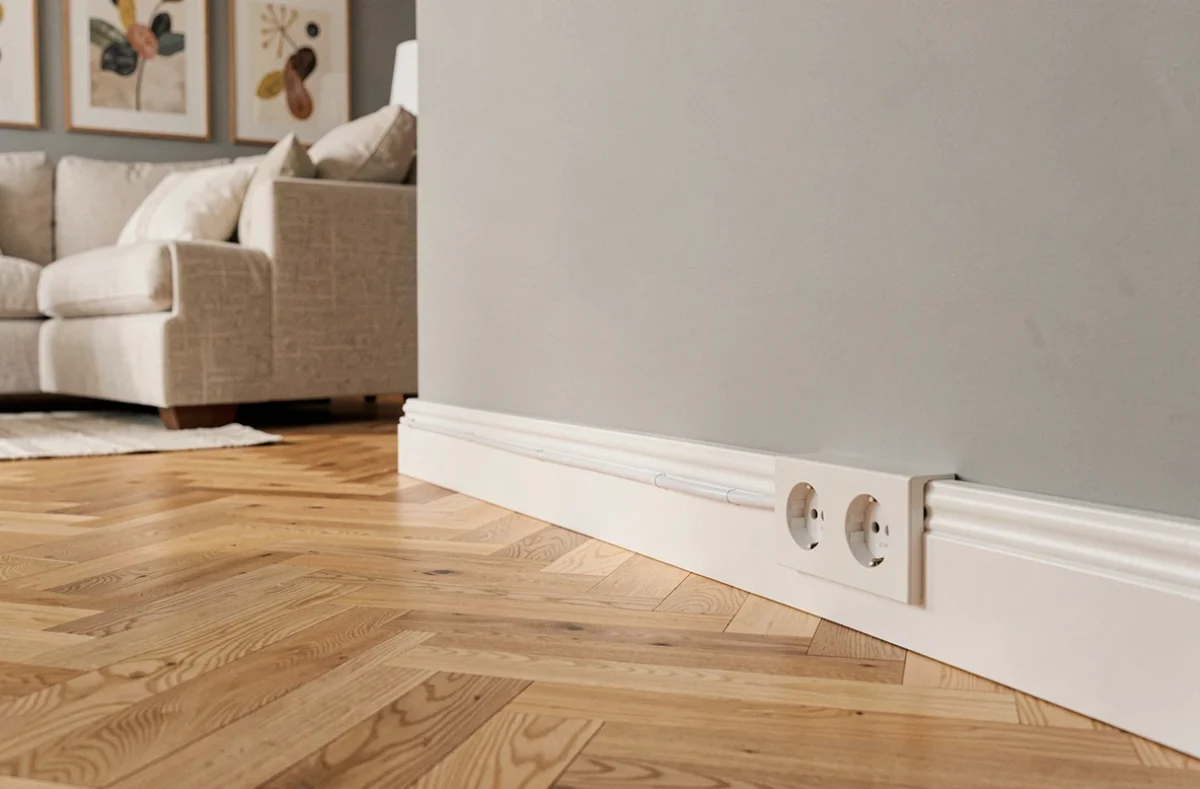 Goulotte électrique blanche en plinthe avec prises intégrées dans un salon rénové au parquet en chêne